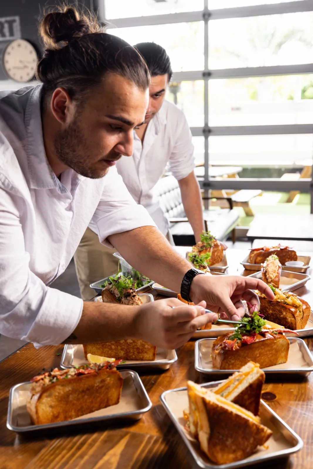 An overhead spread of eleven Lobster Lab rolls and sandwiches plated on individual steel trays, laid out for a group