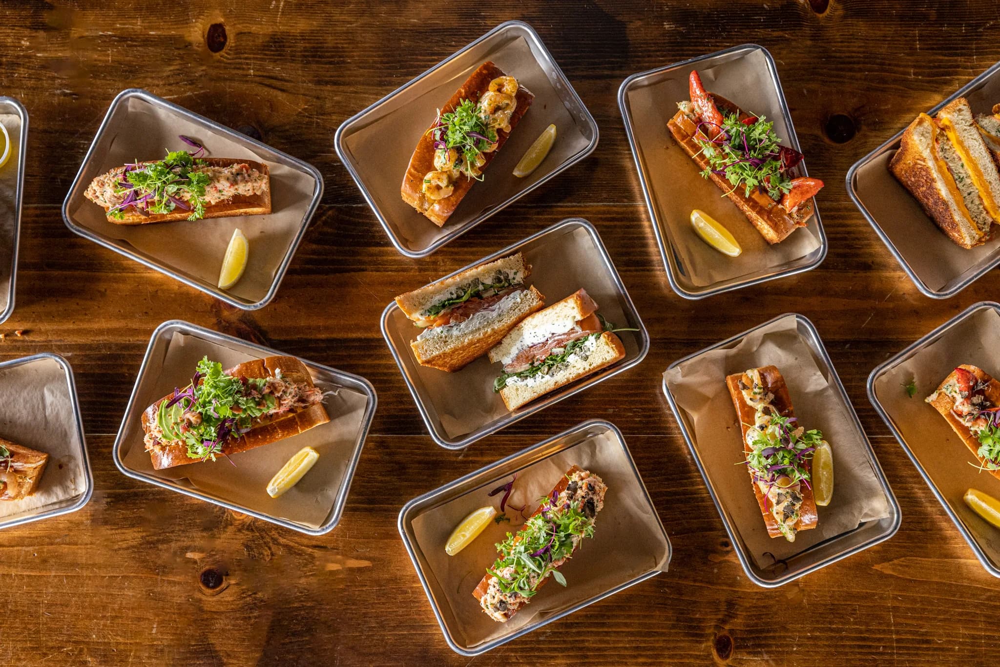 An overhead spread of Lobster Lab lobster rolls, seafood sandwiches and grilled cheese on steel trays